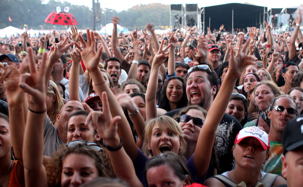 [IMAGE] A large crowd of people with arms raised, smiling at the camera. In the background is an umbrella that looks like a ladybug. The image appears to have been taken of a crowd at a concert during the day time.