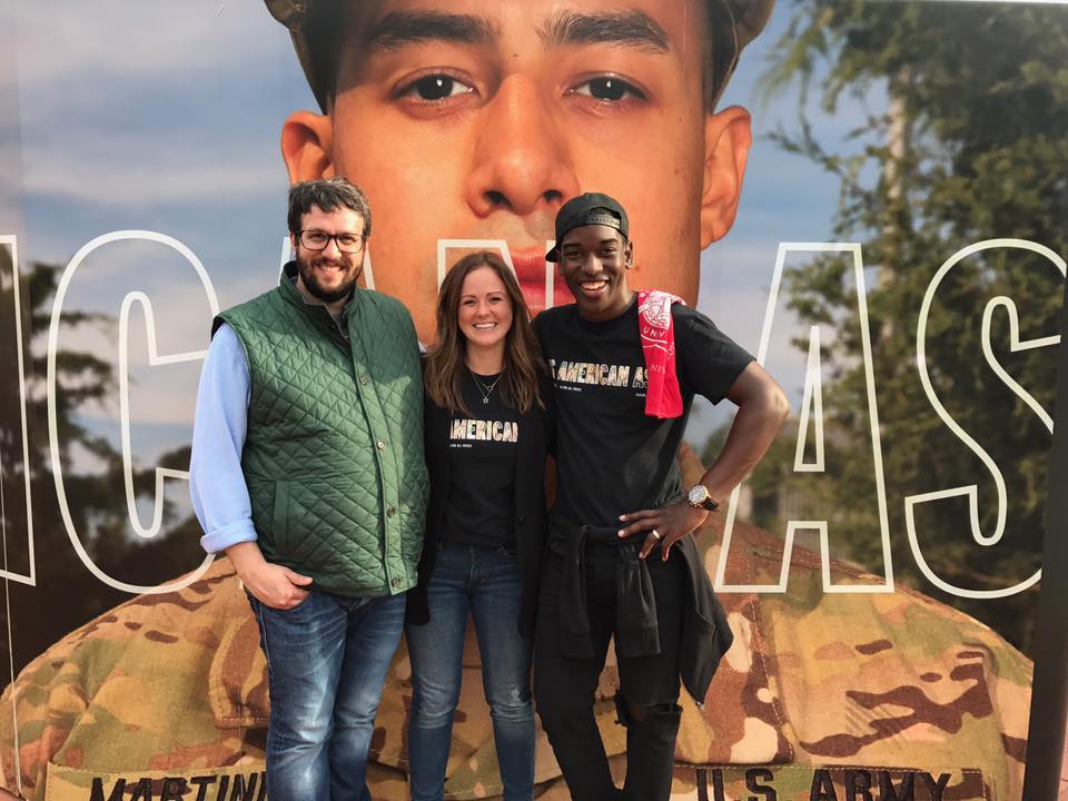 [IMAGE] Three adults - two men, one woman - stand in front of a large image. They have their arms around each other, and they are smiling.