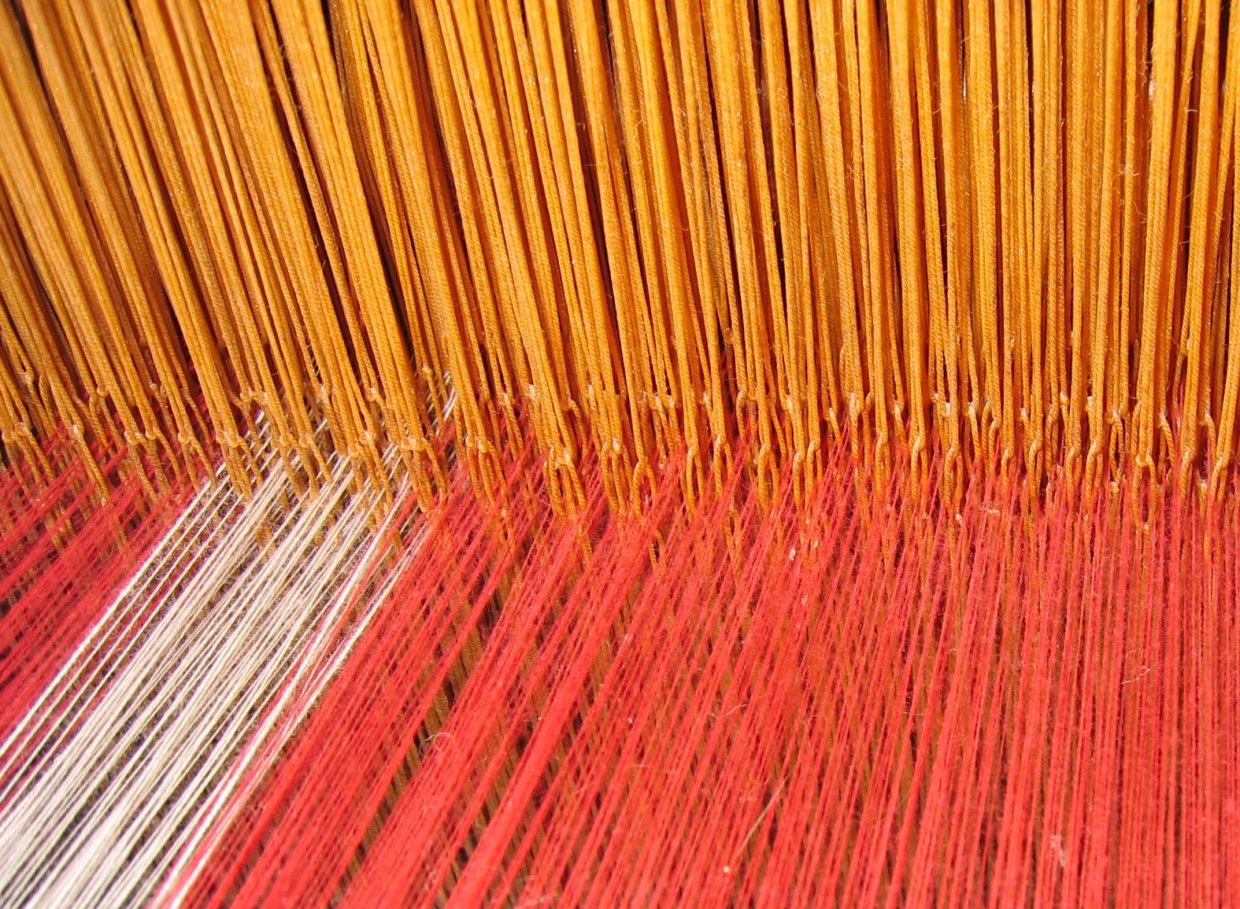 [IMAGE] A close-up of a loom with red, yellow, and white threads combining together.