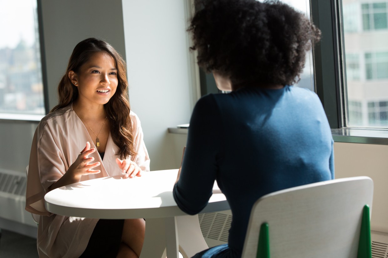 [IMAGE] A woman is sitting at a white table, smiling, talking to another seated women whose back is facing the camera.