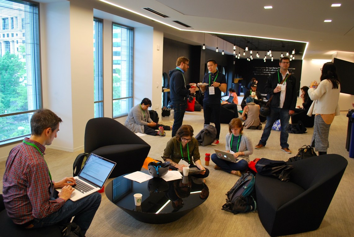 [IMAGE] A group of people, some sitting, some standing, inside a large room. Many are working on laptops. They look to be aged around 20-40 years old. There is a mix of men and women.