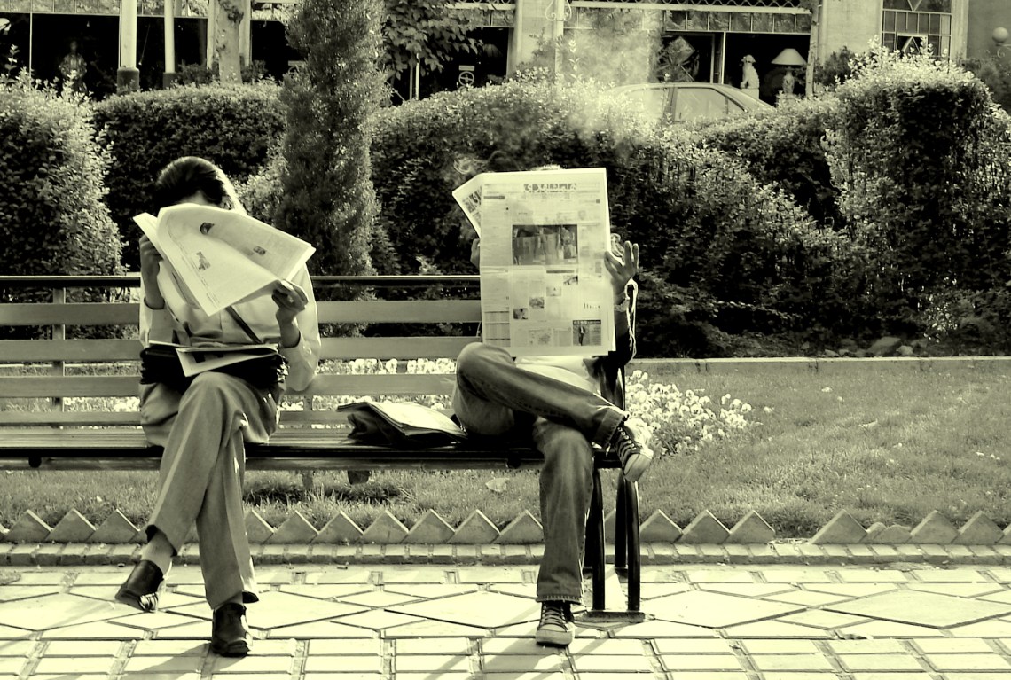 [IMAGE] Two people sat on a part bench reading newspapers. Their faces are obscured.