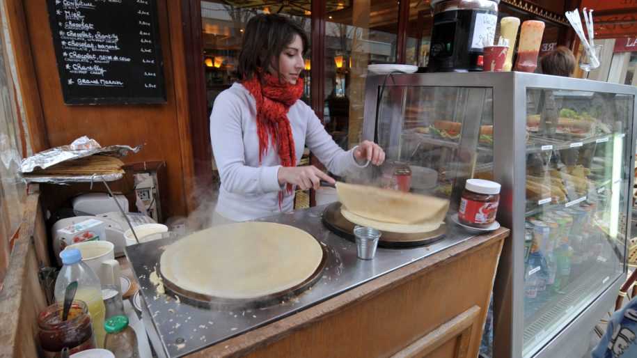 [IMAGE] A light-skinned woman who appears to be in her 20s, wearing a white sweater and bright red scarf, prepares crepes outside of Quasimodo Cafe on the Rue D'arc in Paris